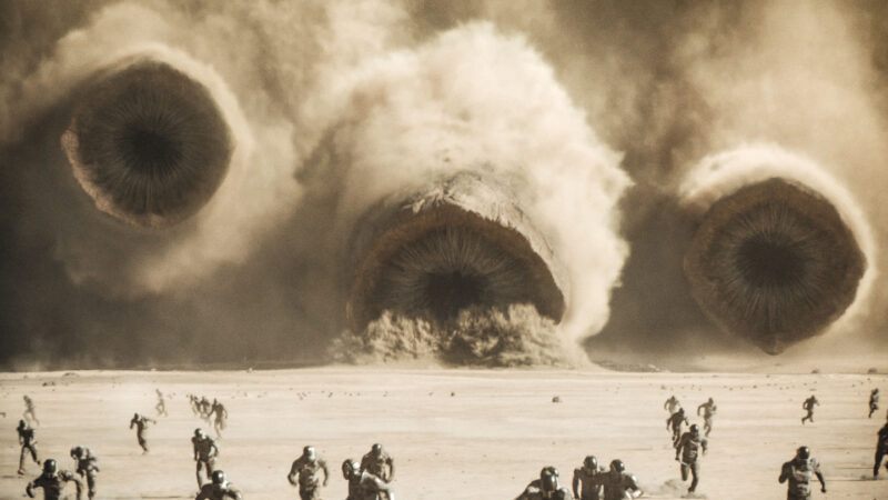 From a cloud of sand, three giant worms emerge. Their circular mouths are open displaying rows of long teeth that curve into their throats. At least two dozen people dressed in dark full-body suits flee toward the camera across an open expanse of sand from the giant worms. The people seem nearly as small as insects in comparison with the huge worms.