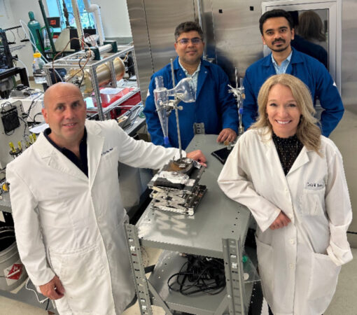 Four researchers stand next to their coronavirus detector placed on a metal table. The two researchers standing next to the device are wearing white lab coats. The two researchers behind the device are wearing blue jackets.