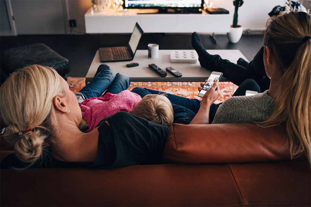 A photo of two adults and a child sitting on a sofa as seen from behind. The bottom edge of a TV can be seen playing a show on the top of the image. There is alos an open laptop on the coffee table in front of the people and at least one adult is also looking at their phone.