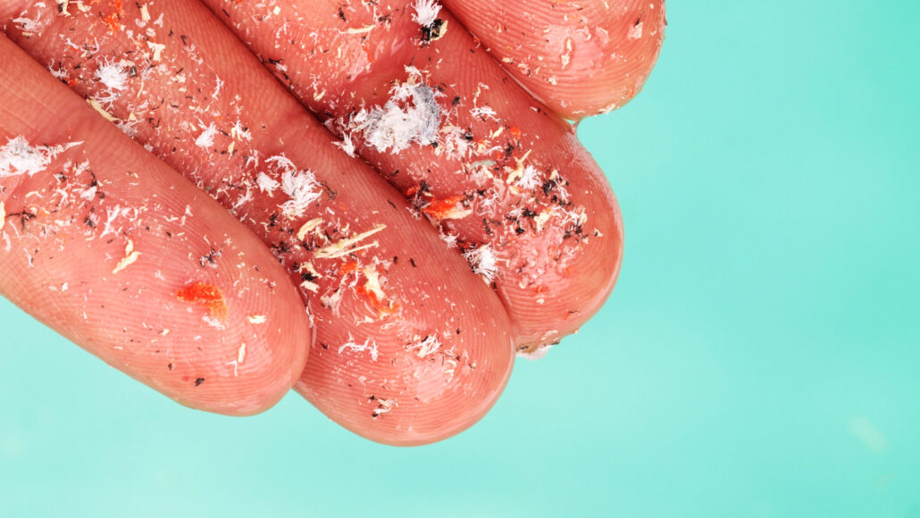 a hand holds four fingertips covered with microplastics close to the camera, in the background is blue-green water