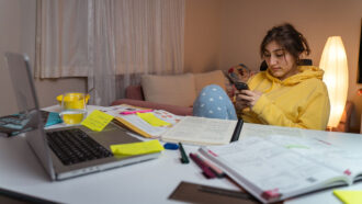 a young lady sitting at a desk and ignoring the study materials and laptop spread out in front of her, instead she is using her phone