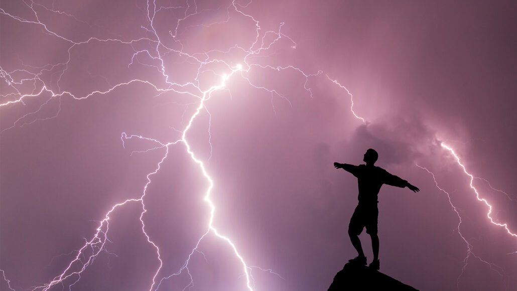 a silhoutte of a person standing on a rocky perch against a purple clouded sky and a lightning strike