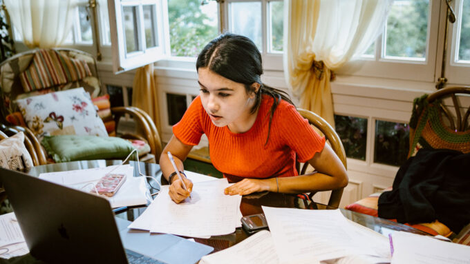 a tween girl hand writes on notebook paper at a desk while reading a laptop screen