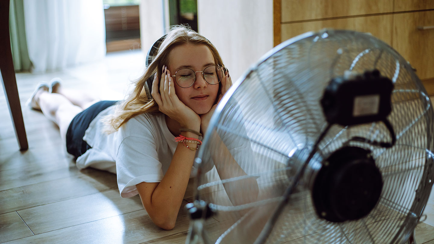 a girl lying on a tile floor in front of a fan, her head is propped up on her hands, she's wearing headphones and her eyes are closed