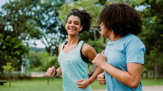 two teens wearing blue workout shirts jog in a park; one is wearing wireless earbuds