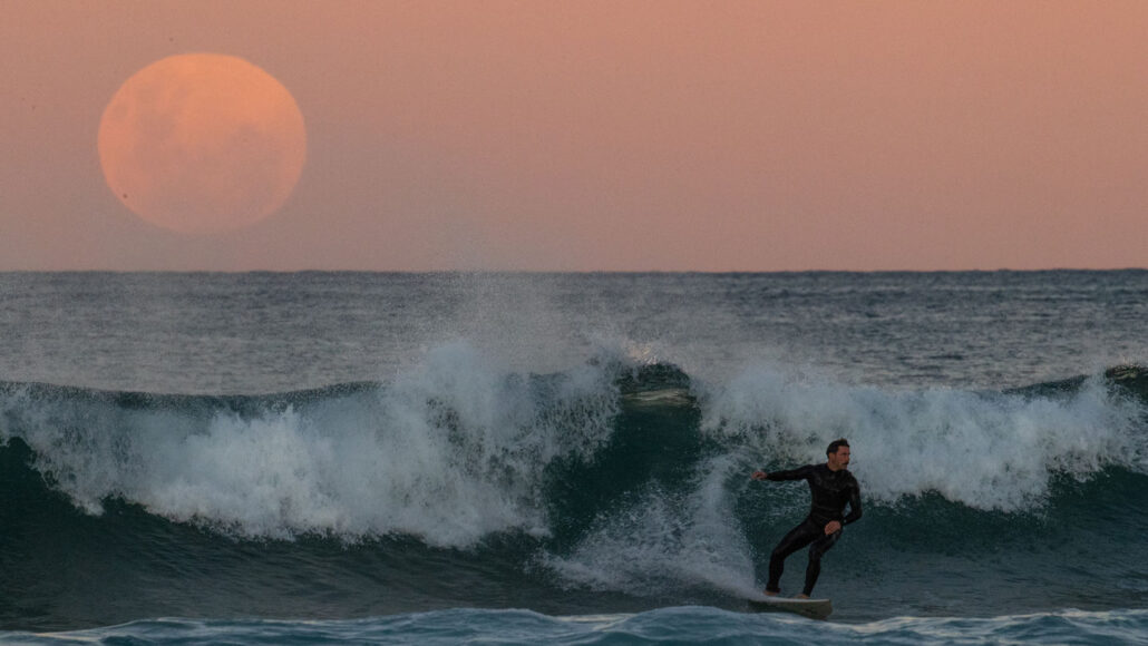 A large reddish full moon is seen over the ocean, with a surfer in the foreground