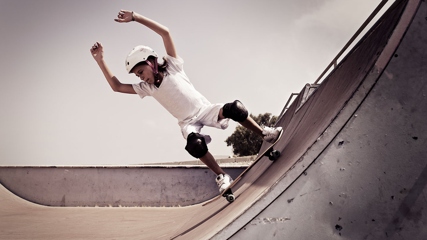 a girl on a skateboard going down a half pipe as seen from the side, she's wearing a white helmet and kneepads