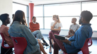 a group of young adults sit in a circle in a brightly lit room while one young woman speaks