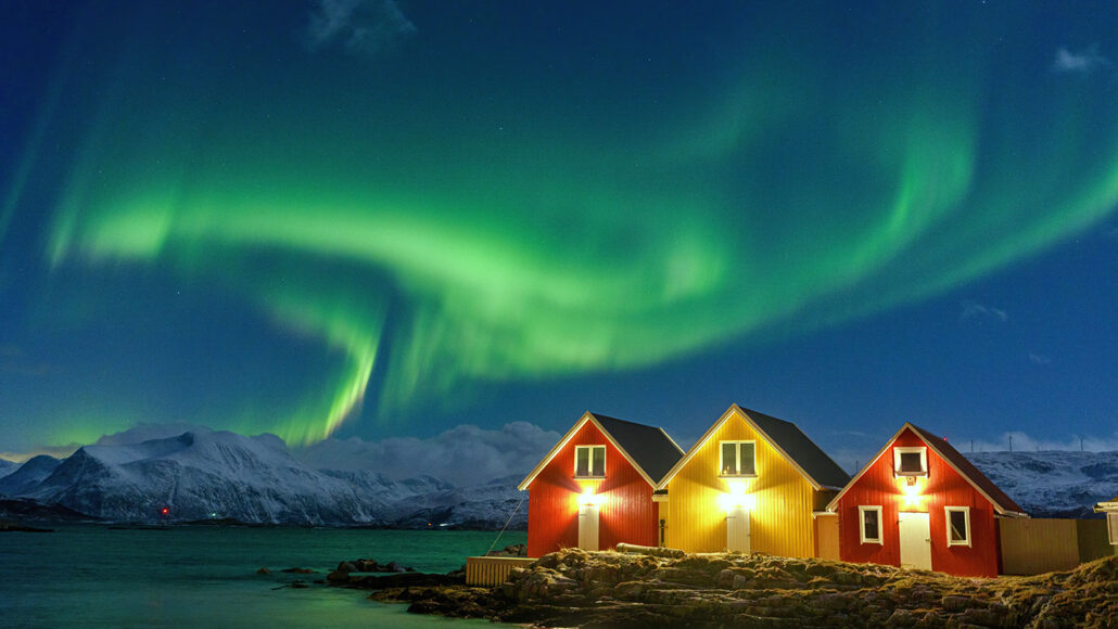 a swirling green aurora borealis fills the night sky above snowcapped mountains, a green-tinted lake and three small cottages