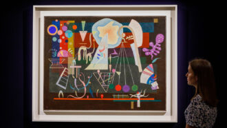 an abstract painting with a medley of different shapes and colors hangs on a black wall; a woman in the corner of the frame gazes at the painting