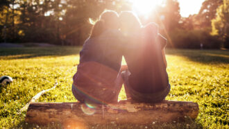 two girls sit on a log with their backs facing the camera, one with her arms around the other