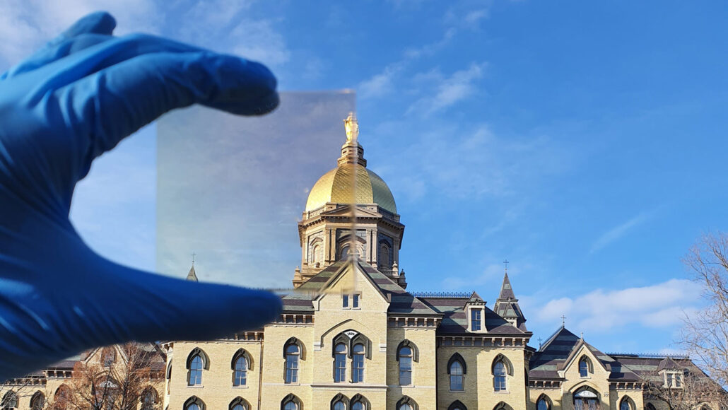 a gloved hand holds a piece of glass up against a building and a blue sky, showing that the glass tints the objects behind it orange