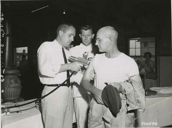 a black and white image showing a man getting a shot for typhus, with a liquid-based jet injector