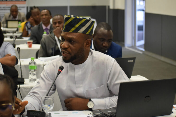 Sunday Agbonika sits at a table with a white cloth as he addresses others at a conference. He is a Black man wearing a white long sleeved shirt and a traditional hat. An open laptop sits before him.