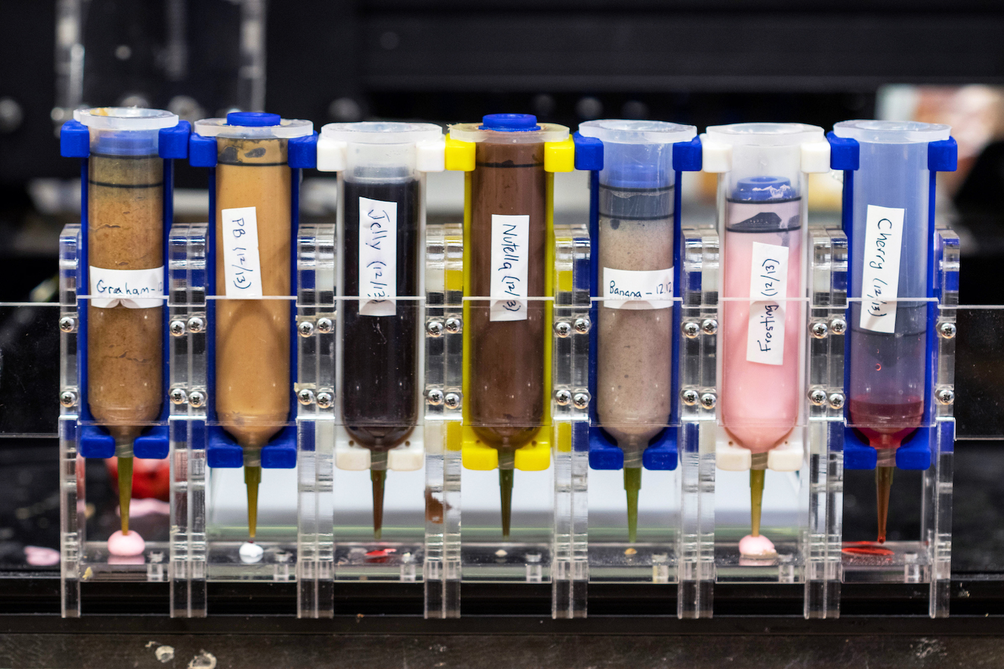 a row of seven transparent upside down nozzles are full of different ingredients, such as Nutella (brown) and frosting (pink)